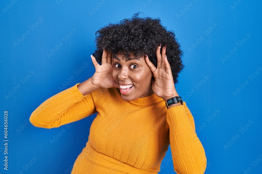 Poster black woman with curly hair standing over blue background smiling cheerful playing peek a boo with h