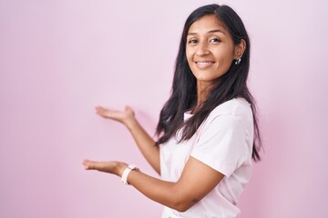 Young hispanic woman standing over pink background inviting to enter smiling natural with open hand