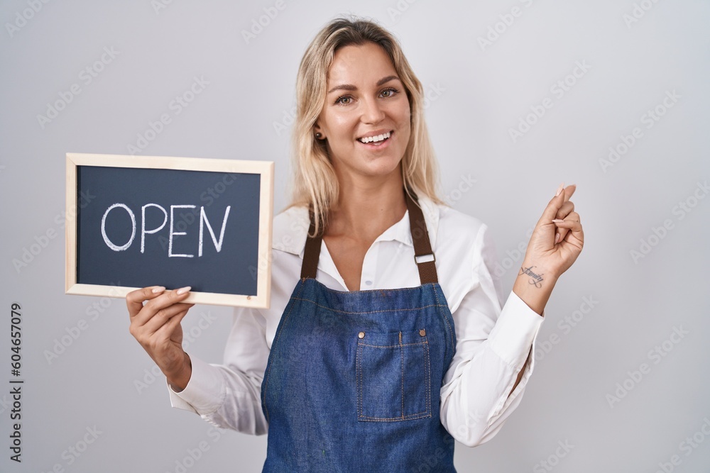 Poster young blonde woman wearing apron holding blackboard with open word smiling happy pointing with hand 