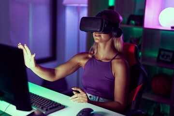 Young blonde woman streamer playing video game using virtual reality glasses at gaming room