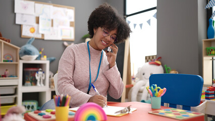 Young african american woman preschool teacher writing on document talking on smartphone at...