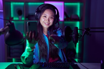 Young asian woman playing video games with smartphone pointing to you and the camera with fingers, smiling positive and cheerful