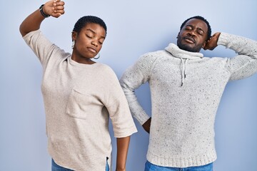 Young african american couple standing over blue background together stretching back, tired and...