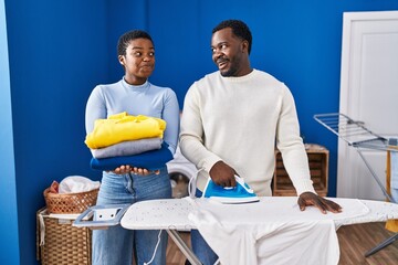 Young african american couple ironing clothes at laundry room smiling looking to the side and staring away thinking.