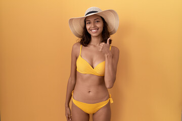 Young hispanic woman wearing bikini and summer hat doing happy thumbs up gesture with hand. approving expression looking at the camera showing success.