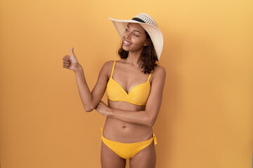 Young hispanic woman wearing bikini and summer hat looking proud, smiling doing thumbs up gesture to the side