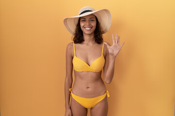 Young hispanic woman wearing bikini and summer hat showing and pointing up with fingers number five while smiling confident and happy.