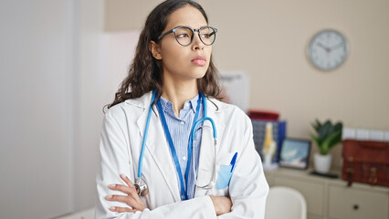 Young beautiful hispanic woman doctor standing with serious expression and crossed arms at clinic