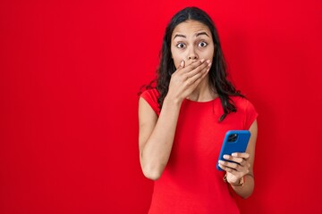 Young brazilian woman using smartphone over red background shocked covering mouth with hands for mistake. secret concept.