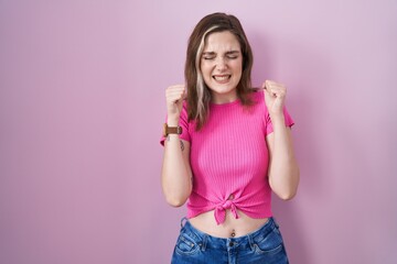 Blonde caucasian woman standing over pink background excited for success with arms raised and eyes closed celebrating victory smiling. winner concept.