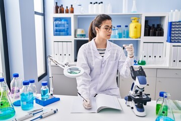 Young beautiful hispanic woman scientist holding sample writing on notebook at laboratory