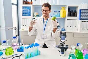 Young hispanic man working at scientist laboratory holding apple winking looking at the camera with...