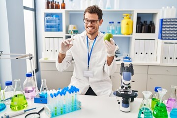 Young hispanic man working at scientist laboratory holding apple winking looking at the camera with sexy expression, cheerful and happy face.