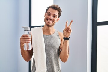Young hispanic man wearing sportswear drinking water smiling with happy face winking at the camera...