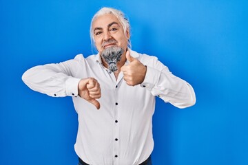 Fototapeta premium Middle age man with grey hair standing over blue background doing thumbs up and down, disagreement and agreement expression. crazy conflict