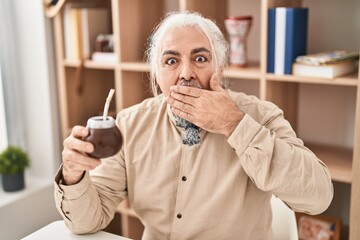 Middle age man with grey hair drinking mate infusion covering mouth with hand, shocked and afraid for mistake. surprised expression