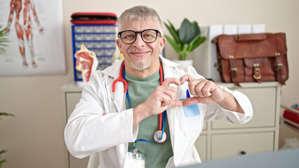 Middle age grey-haired man doctor smiling doing heart gesture with hands at clinic