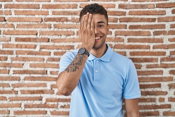 Brazilian young man standing over brick wall covering one eye with hand, confident smile on face and surprise emotion.