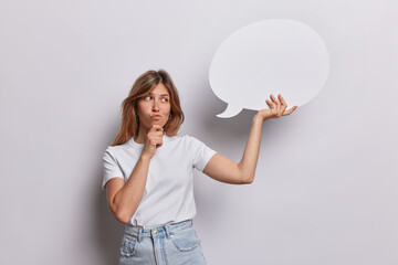 Pensive young European woman holds chin and looks doubtful holds blank speech communication bubble...