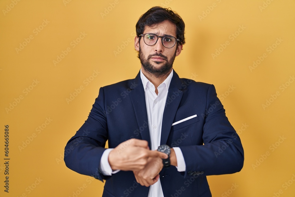 Poster handsome latin man standing over yellow background in hurry pointing to watch time, impatience, upse