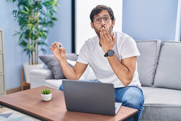 Handsome latin man holding virtual currency bitcoin using laptop covering mouth with hand, shocked...