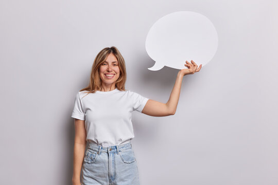 Great idea concept. Smiling young European woman smiles broadly holds empty speech bubble expresses her opinion and feedback dressed in casual t shirt and jeans isolated over white background