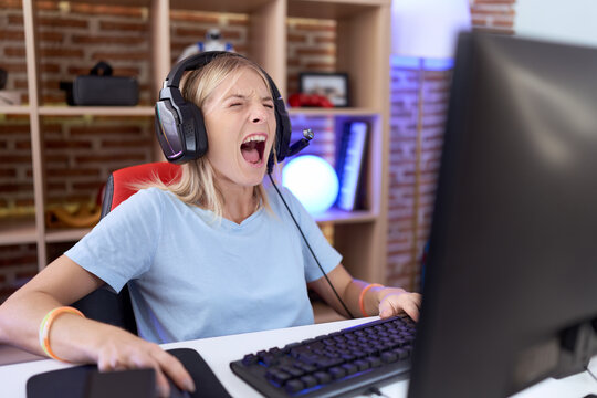 Young caucasian woman playing video games wearing headphones angry and mad screaming frustrated and furious, shouting with anger. rage and aggressive concept.
