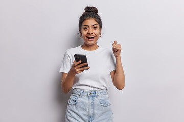 Happy cheerful Indian girl with dark hair combed in bun clenches fist celebrates success holds smartphone dressed in casual basic t shirt jeans isolated over white wall receives message about reward - 604665660