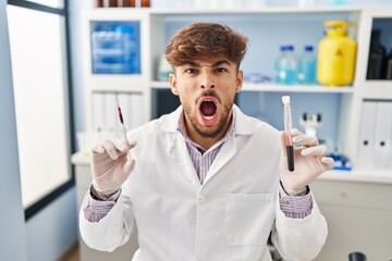 Arab man with beard working at scientist laboratory holding blood sample angry and mad screaming frustrated and furious, shouting with anger. rage and aggressive concept.