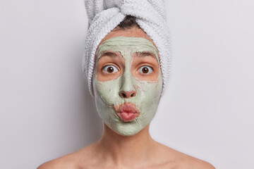 Headshot of surprised young woman applies beauty cosmetic mask undergoes facial skin care stands shirtless indoor wears bath towel on head isolated over white background. Pampering procedures