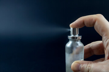 Selective blur on fingers pushing the spray of a small perfume bottle, spreading in aerosols droplets of fragrance with scent of a high class eau de toilette parfum, in front of a black background