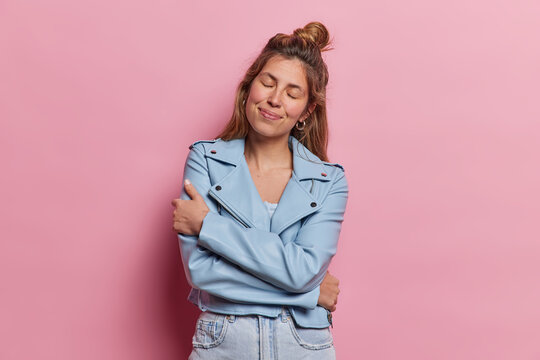 Comfort and tenderness concept. Charming young European woman has eyes closed ebraces herself feels cozy dressed in fashionable jacket and jeans isolated over pink background. I love myself.