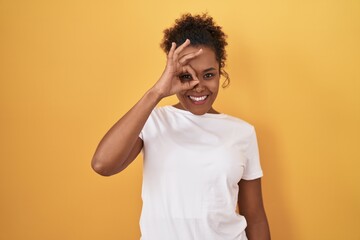 Young hispanic woman with curly hair standing over yellow background doing ok gesture with hand smiling, eye looking through fingers with happy face.