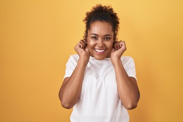 Young hispanic woman with curly hair standing over yellow background smiling pulling ears with...