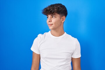 Hispanic teenager standing over blue background looking away to side with smile on face, natural...