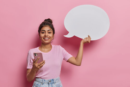 Horizontal shot of cheerful Indian woman smiles toothily being in good mood uses smartphone for chatting online holds white speech bubble for placing your idea dressed casually isolated over pink wall