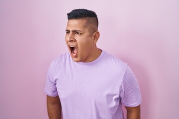 Young hispanic man standing over pink background angry and mad screaming frustrated and furious,...