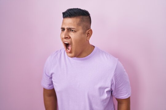 Young hispanic man standing over pink background angry and mad screaming frustrated and furious, shouting with anger. rage and aggressive concept.