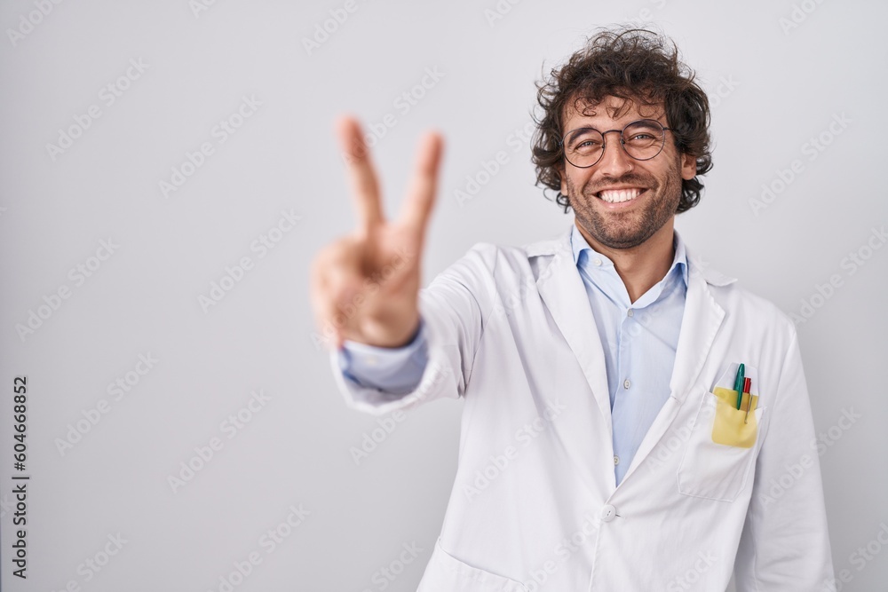 Sticker hispanic young man wearing doctor uniform smiling looking to the camera showing fingers doing victor