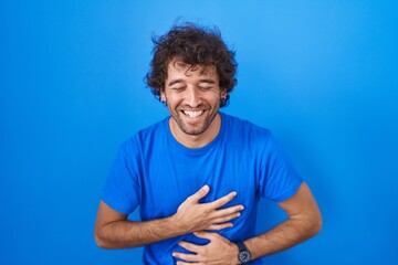 Fototapeta na wymiar Hispanic young man standing over blue background smiling and laughing hard out loud because funny crazy joke with hands on body.