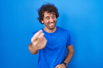 Hispanic young man standing over blue background beckoning come here gesture with hand inviting welcoming happy and smiling