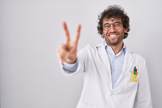 Hispanic young man wearing doctor uniform smiling looking to the camera showing fingers doing victory sign. number two.