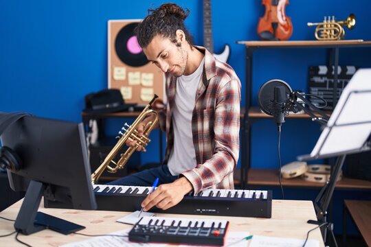 Young hispanic man musician composing song holding trumpet at music studio