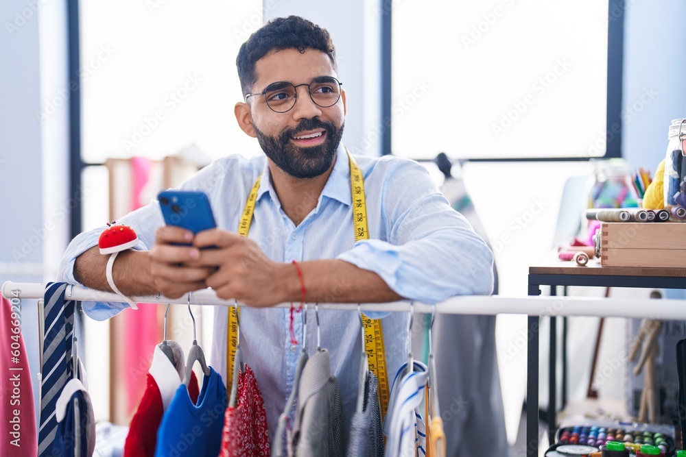 Poster young arab man tailor using smartphone leaning on clothes rack at tailor shop