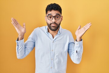 Hispanic man with beard standing over yellow background clueless and confused expression with arms...