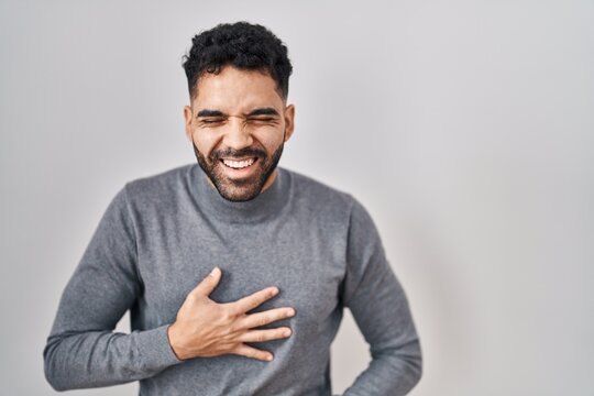 Hispanic man with beard standing over white background smiling and laughing hard out loud because funny crazy joke with hands on body.