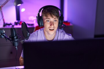 Caucasian blond man playing video games scared and amazed with open mouth for surprise, disbelief...