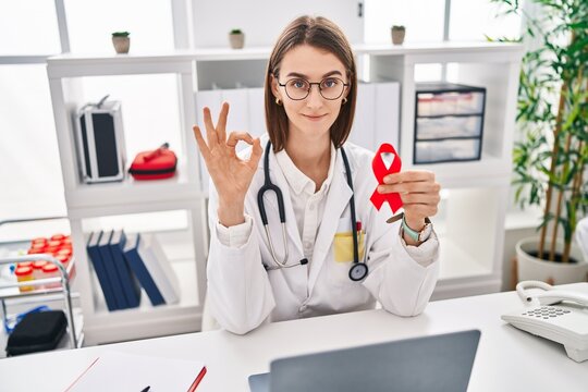 Young caucasian doctor woman holding support red ribbon doing ok sign with fingers, smiling friendly gesturing excellent symbol
