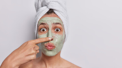 Horizontal shot of surprised young woman applies natural beauty mask on face for skin care touches nose wears bath towel on head isolated over white background blank space for your advertising content