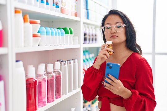 Young chinese woman customer using smartphone holding medicine bottle at pharmacy